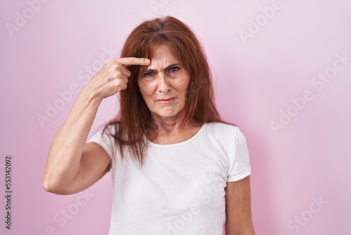 Middle age woman standing over pink background pointing unhappy to pimple on forehead, ugly infection of blackhead. acne and skin problem