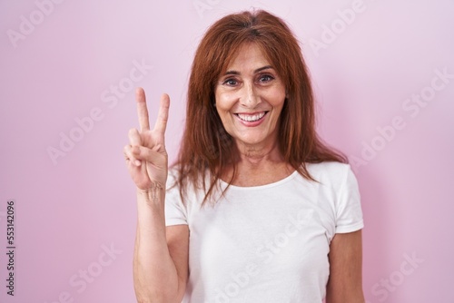 Middle age woman standing over pink background showing and pointing up with fingers number two while smiling confident and happy.
