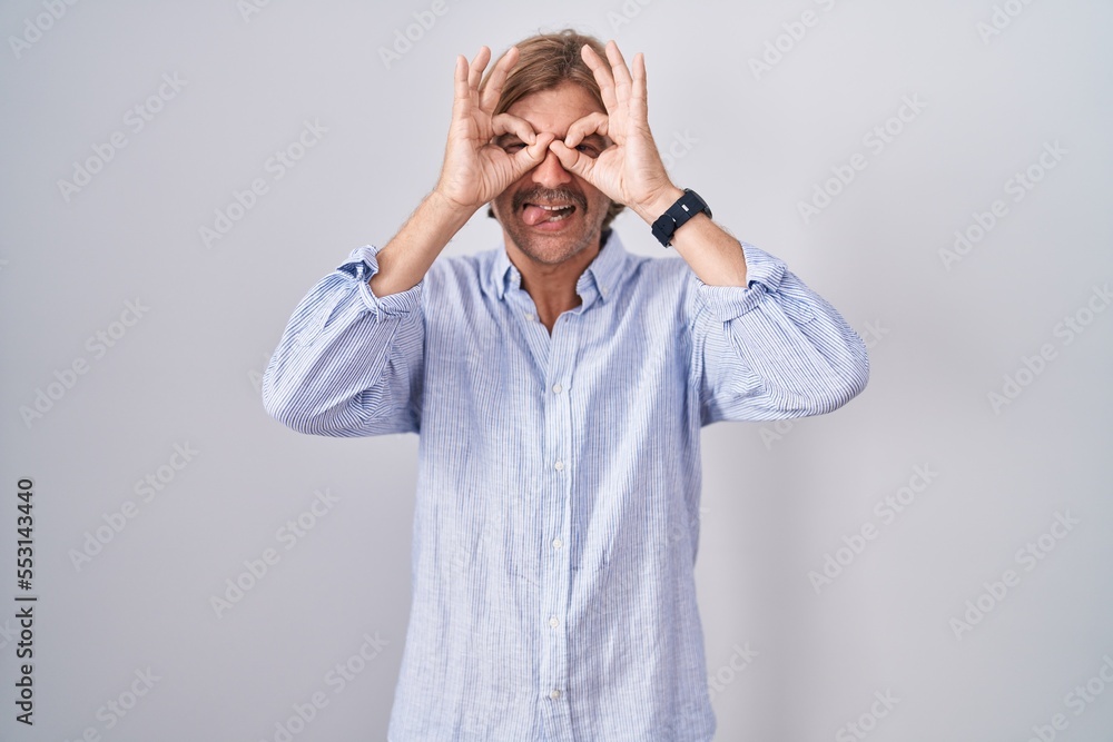 Caucasian man with mustache standing over white background doing ok gesture like binoculars sticking tongue out, eyes looking through fingers. crazy expression. Caucasian man with mustache standing over white background doing ok gesture like binoculars sticking tongue out, eyes looking through fingers. crazy expression.