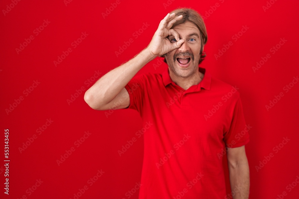 Caucasian man with mustache standing over red background doing ok gesture with hand smiling, eye looking through fingers with happy face. Caucasian man with mustache standing over red background doing ok gesture with hand smiling, eye looking through fingers with happy face.