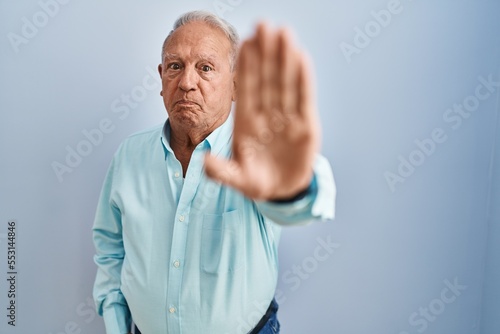 Senior man with grey hair standing over blue background doing stop sing with palm of the hand. warning expression with negative and serious gesture on the face.