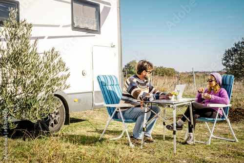 Happy tourist couple enjoy outdoor leisure activity sitting on the table with camper van in background. Alternative house lifestyle and nomadic vacation off grid. Travel with van man and woman people