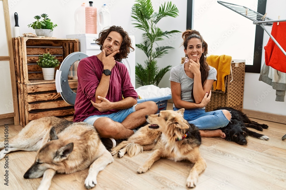 Young hispanic couple doing laundry with dogs looking confident at the camera smiling with crossed arms and hand raised on chin. thinking positive.