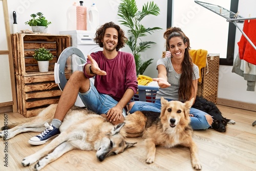 Young hispanic couple doing laundry with dogs smiling cheerful offering palm hand giving assistance and acceptance.