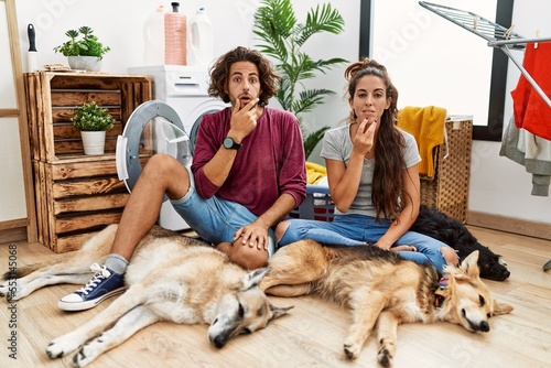 Photography Young hispanic couple doing laundry with dogs looking fascinated with disbelief,