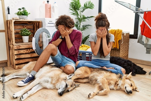 Sticker Young hispanic couple doing laundry with dogs with sad expression covering face with hands while crying