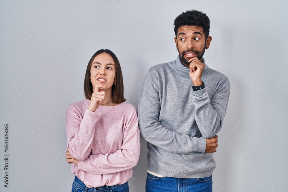 Young hispanic couple standing together thinking worried about a question, concerned and nervous with hand on chin Young hispanic couple standing together thinking worried about a question, concerned and nervous with hand on chin