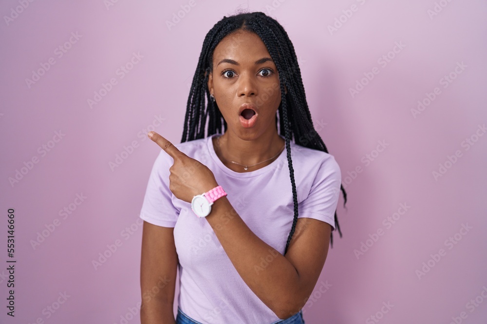 African american woman with braids standing over pink background surprised pointing with finger to the side, open mouth amazed expression. African american woman with braids standing over pink background surprised pointing with finger to the side, open mouth amazed expression.
