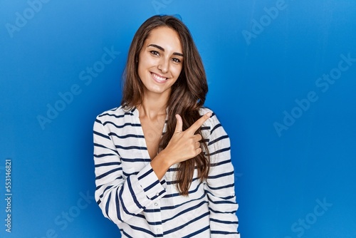 Young hispanic woman standing over blue isolated background cheerful with a smile on face pointing with hand and finger up to the side with happy and natural expression
