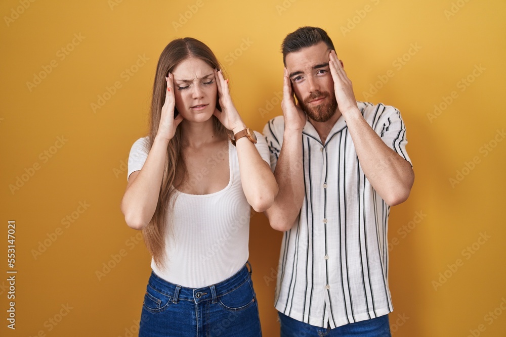Young couple standing over yellow background with hand on head, headache because stress. suffering migraine. Young couple standing over yellow background with hand on head, headache because stress. suffering migraine.