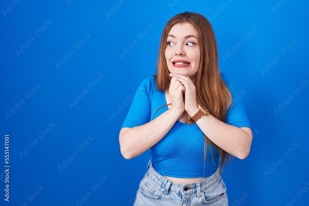 Redhead woman standing over blue background laughing nervous and excited with hands on chin looking to the side Redhead woman standing over blue background laughing nervous and excited with hands on chin looking to the side