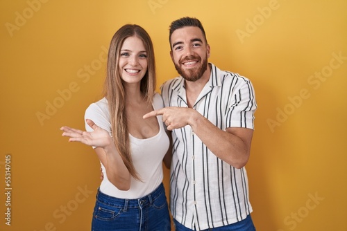 Young couple standing over yellow background amazed and smiling to the camera while presenting with hand and pointing with finger.