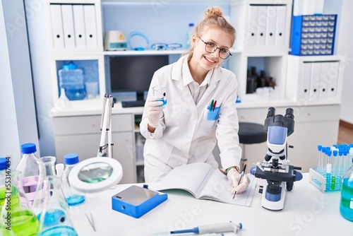 Young blonde woman scientist writing on notebook holding test tube at laboratory