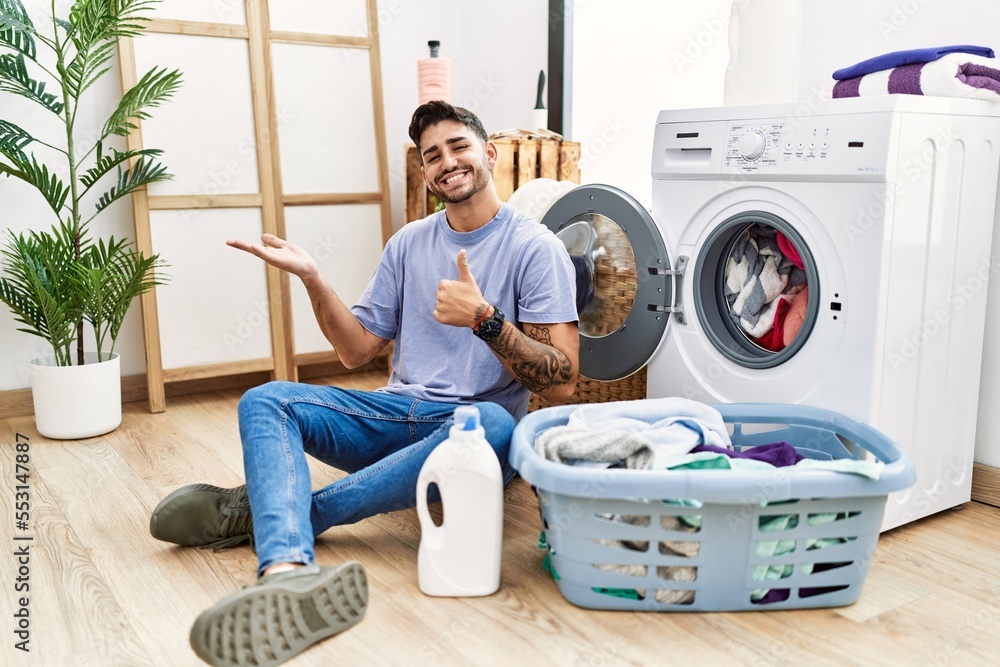 Young hispanic man putting dirty laundry into washing machine showing palm hand and doing ok gesture with thumbs up, smiling happy and cheerful Young hispanic man putting dirty laundry into washing machine showing palm hand and doing ok gesture with thumbs up, smiling happy and cheerful
