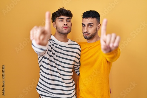 Young hispanic gay couple standing over yellow background pointing with finger up and angry expression  showing no gesture