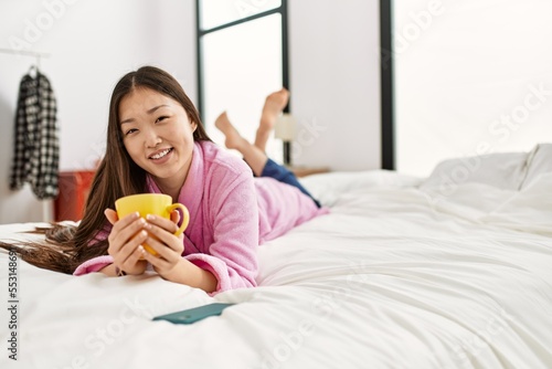 Young chinese girl drinking coffee lying on the bed at bedroom.