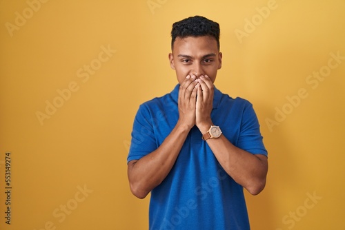 Fotografie Young hispanic man standing over yellow background laughing and embarrassed gigg