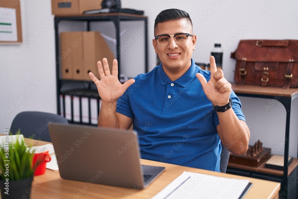 Young hispanic man working at the office with laptop showing and pointing up with fingers number seven while smiling confident and happy.