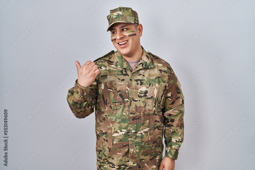 Hispanic young man wearing camouflage army uniform smiling with happy face looking and pointing to the side with thumb up. Hispanic young man wearing camouflage army uniform smiling with happy face looking and pointing to the side with thumb up.
