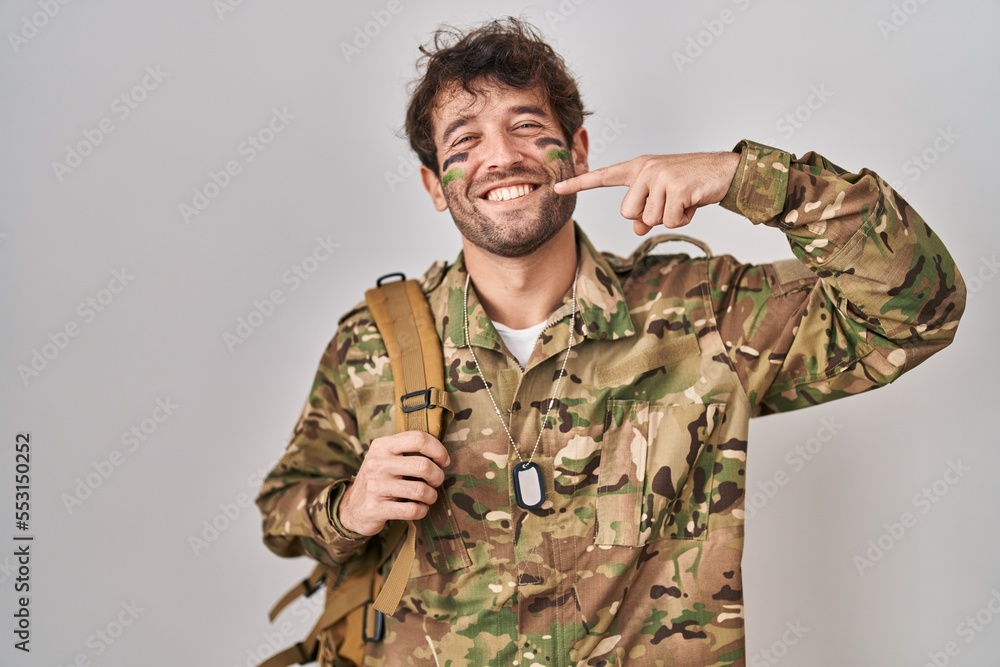 Hispanic young man wearing camouflage army uniform smiling cheerful showing and pointing with fingers teeth and mouth. dental health concept.