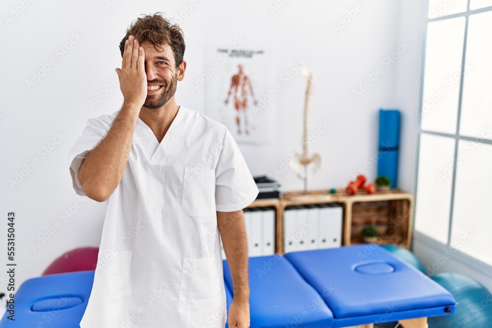 Young handsome physiotherapist man working at pain recovery clinic covering one eye with hand, confident smile on face and surprise emotion. Young handsome physiotherapist man working at pain recovery clinic covering one eye with hand, confident smile on face and surprise emotion.