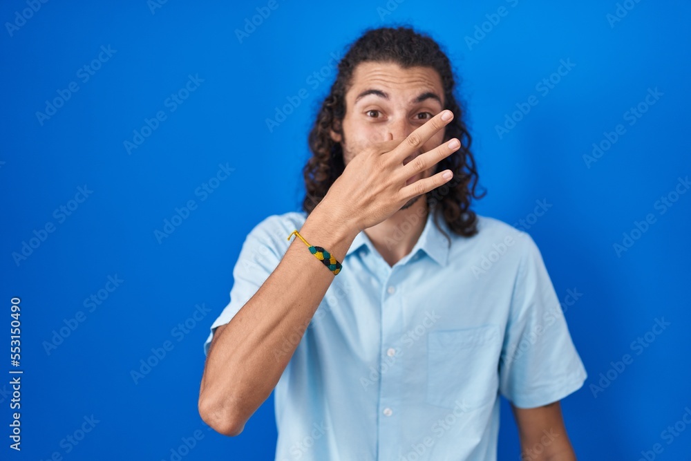 Young hispanic man standing over blue background smelling something stinky and disgusting, intolerable smell, holding breath with fingers on nose. bad smell Young hispanic man standing over blue background smelling something stinky and disgusting, intolerable smell, holding breath with fingers on nose. bad smell