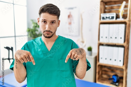 Young physiotherapist man working at pain recovery clinic pointing down looking sad and upset, indicating direction with fingers, unhappy and depressed.