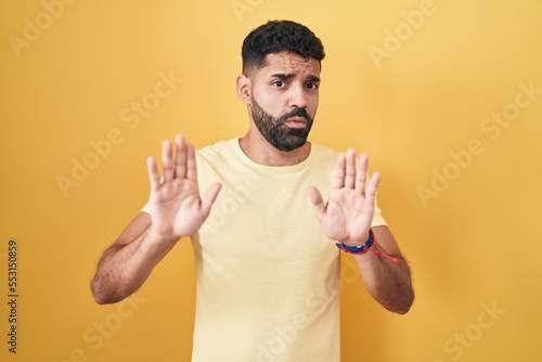 Hispanic man with beard standing over yellow background moving away hands palms showing refusal and denial with afraid and disgusting expression. stop and forbidden.