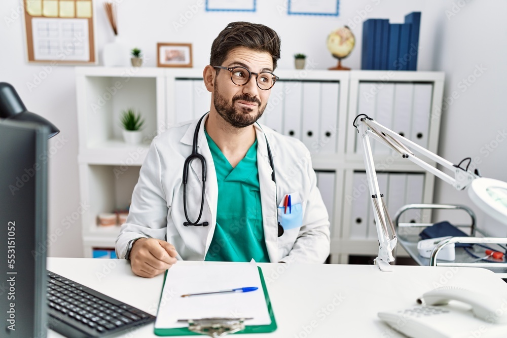 Young man with beard wearing doctor uniform and stethoscope at the clinic smiling looking to the side and staring away thinking. Young man with beard wearing doctor uniform and stethoscope at the clinic smiling looking to the side and staring away thinking.
