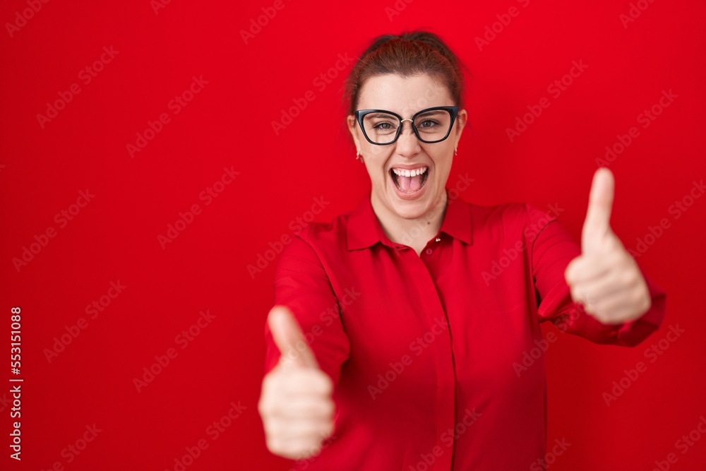 Young hispanic woman with red hair standing over red background approving doing positive gesture with hand, thumbs up smiling and happy for success. winner gesture. Young hispanic woman with red hair standing over red background approving doing positive gesture with hand, thumbs up smiling and happy for success. winner gesture.