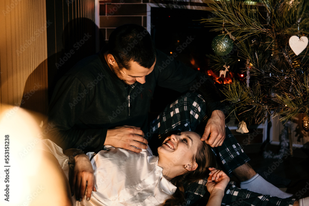 Merry Christmas. Young couple in love celebrating Christmas or new year eve at home. Romantic couple in pajamas sitting and hugging near christmas tree bokeh lights. Merry Christmas. Young couple in love celebrating Christmas or new year eve at home. Romantic couple in pajamas sitting and hugging near christmas tree bokeh lights.