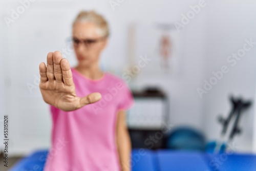 Middle age blonde woman at pain recovery clinic doing stop sing with palm of the hand. warning expression with negative and serious gesture on the face.