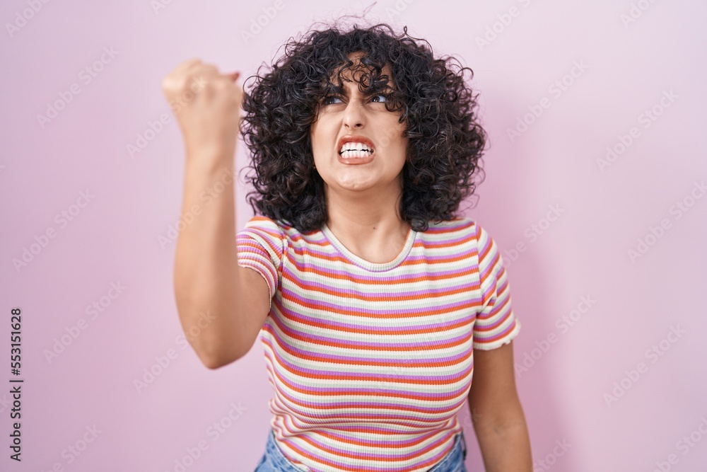 Young middle east woman standing over pink background angry and mad raising fist frustrated and furious while shouting with anger. rage and aggressive concept.