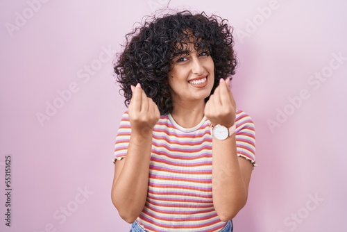 Young middle east woman standing over pink background doing money gesture with hands, asking for salary payment, millionaire business