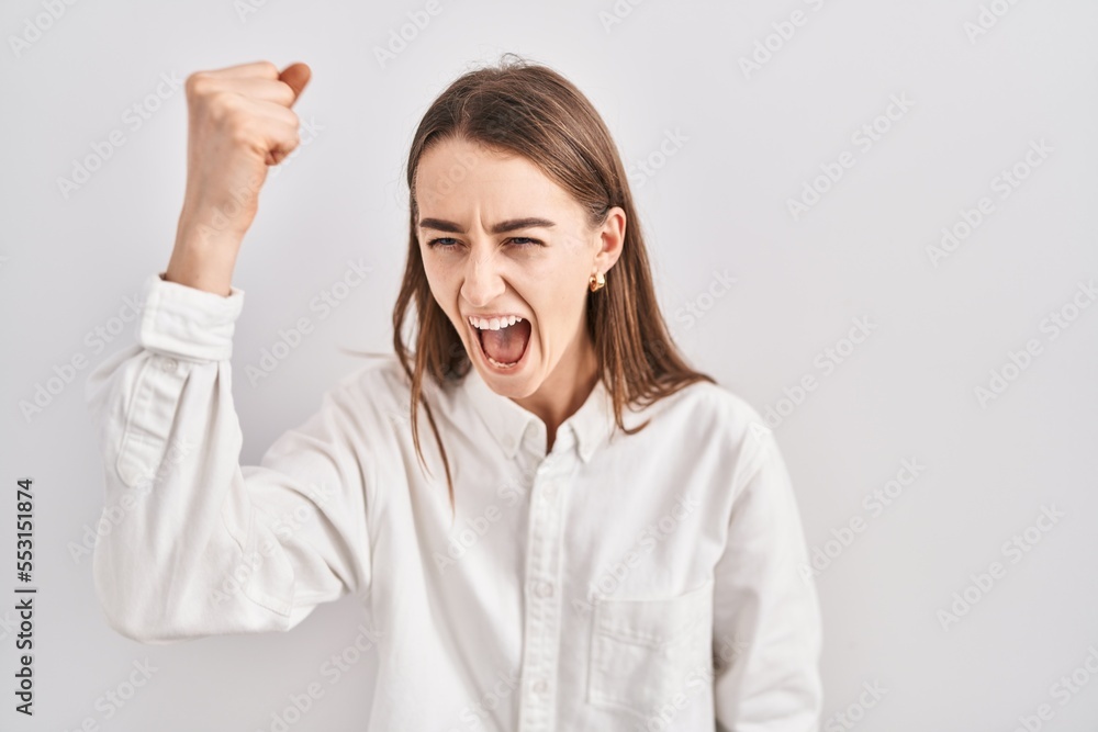 Young caucasian woman standing over isolated background angry and mad raising fist frustrated and furious while shouting with anger. rage and aggressive concept. Young caucasian woman standing over isolated background angry and mad raising fist frustrated and furious while shouting with anger. rage and aggressive concept.