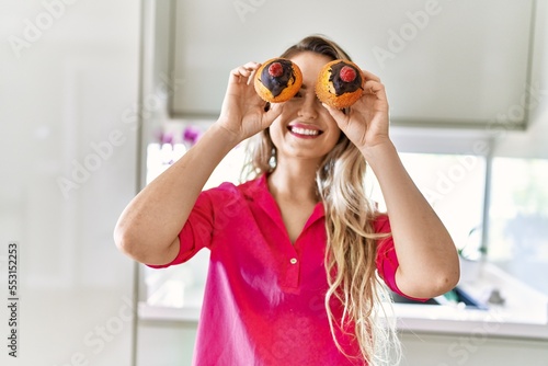 Young woman smiling confident holding cupcakes over eyes at kitchen