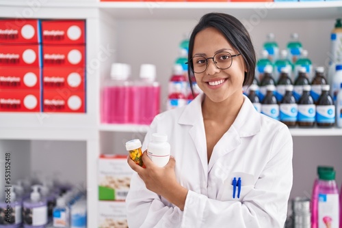 Young hispanic woman pharmacist holding pills bottles standing with arms crossed gesture at pharmacy