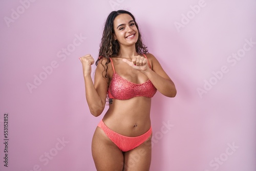 Young hispanic woman wearing lingerie over pink background pointing to the back behind with hand and thumbs up, smiling confident