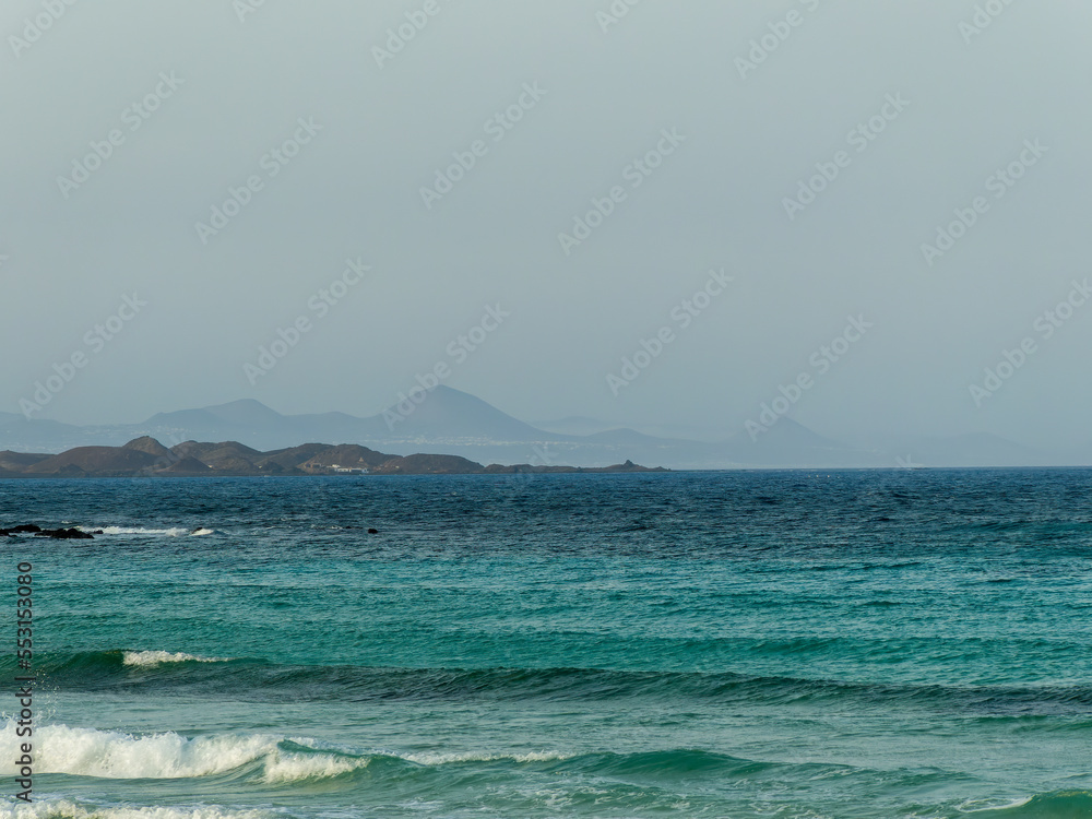 View of Corralejo Bay with open sea, waves and mountains shrouded in mist in the background, Spain