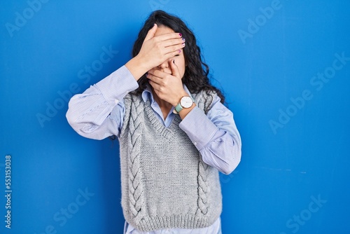 Young brunette woman standing over blue background covering eyes and mouth with hands, surprised and shocked. hiding emotion