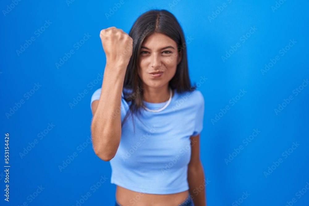 Brunette young woman standing over blue background angry and mad raising fist frustrated and furious while shouting with anger. rage and aggressive concept. Brunette young woman standing over blue background angry and mad raising fist frustrated and furious while shouting with anger. rage and aggressive concept.