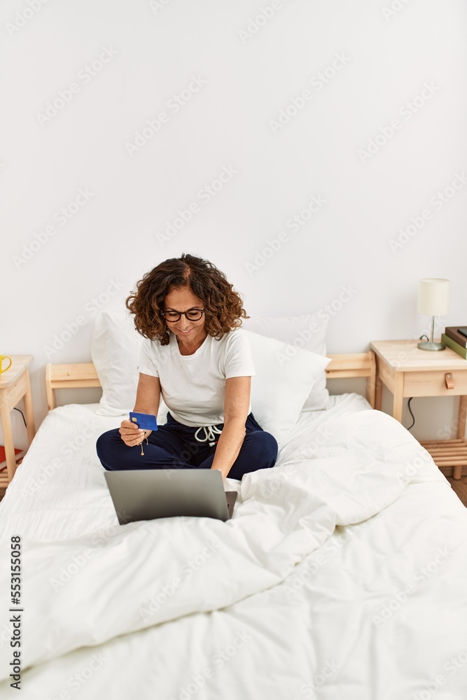 Middle age hispanic woman using laptop holding credit card at bedroom