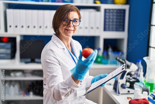 Middle age woman scientist holding apple reading report at laboratory