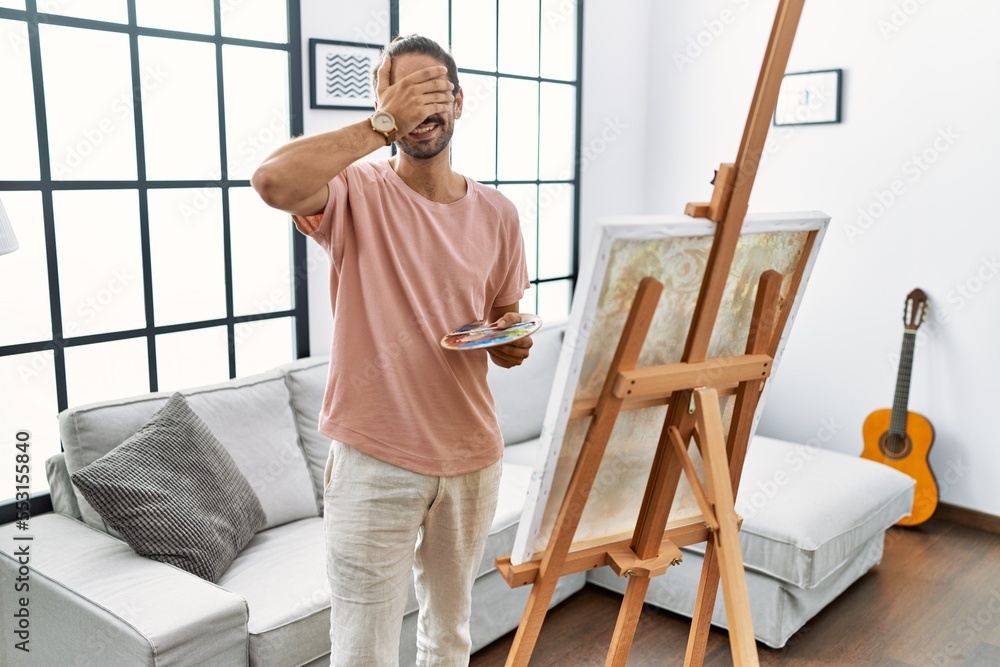 Young hispanic man with beard painting on canvas at home smiling and laughing with hand on face covering eyes for surprise. blind concept. Young hispanic man with beard painting on canvas at home smiling and laughing with hand on face covering eyes for surprise. blind concept.