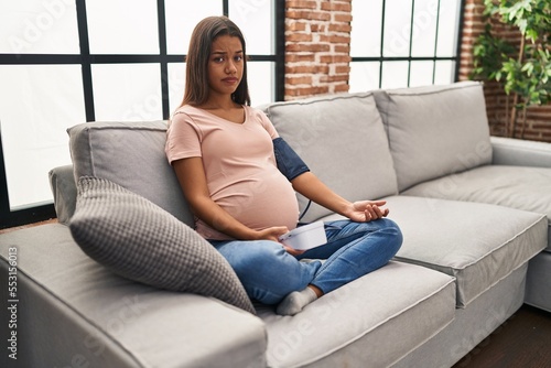 Young pregnant woman using blood pressure monitor sitting on the sofa skeptic and nervous, frowning upset because of problem. negative person.