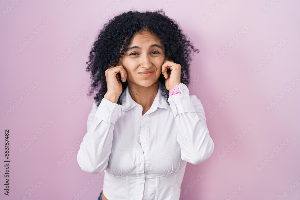 Hispanic woman with curly hair standing over pink background covering ears with fingers with annoyed expression for the noise of loud music. deaf concept. Hispanic woman with curly hair standing over pink background covering ears with fingers with annoyed expression for the noise of loud music. deaf concept.