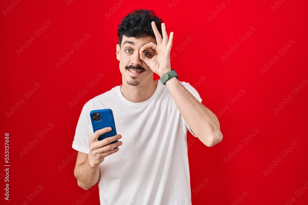 Hispanic man using smartphone over red background doing ok gesture with hand smiling, eye looking through fingers with happy face. Hispanic man using smartphone over red background doing ok gesture with hand smiling, eye looking through fingers with happy face.