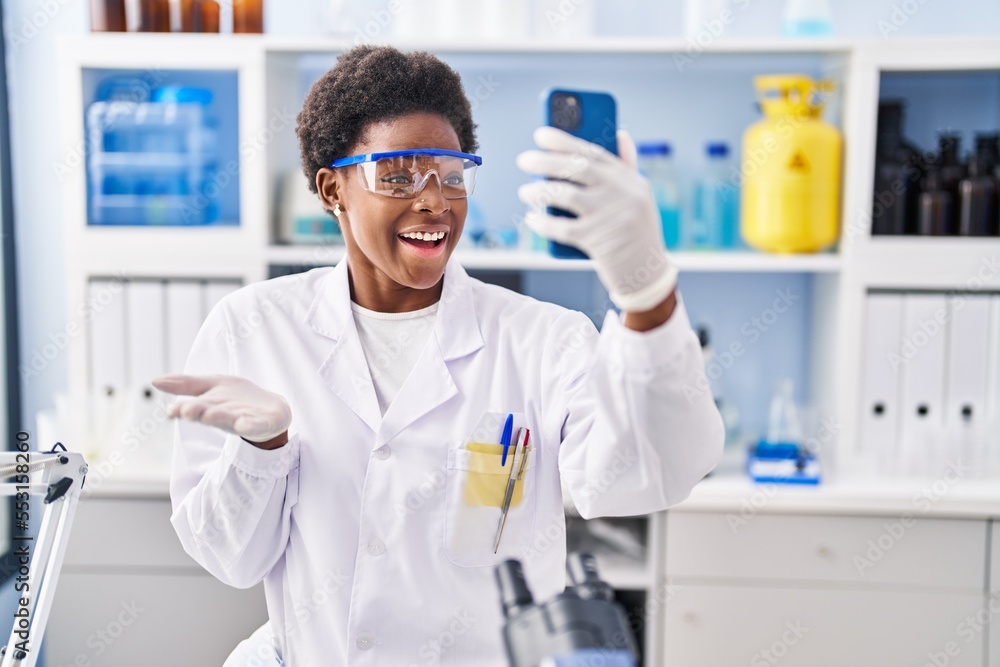 African american woman working at scientist laboratory doing video call with smartphone celebrating achievement with happy smile and winner expression with raised hand African american woman working at scientist laboratory doing video call with smartphone celebrating achievement with happy smile and winner expression with raised hand