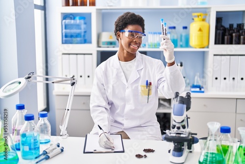 African american woman wearing scientist uniform write on clipboard holding test tubes at laboratory