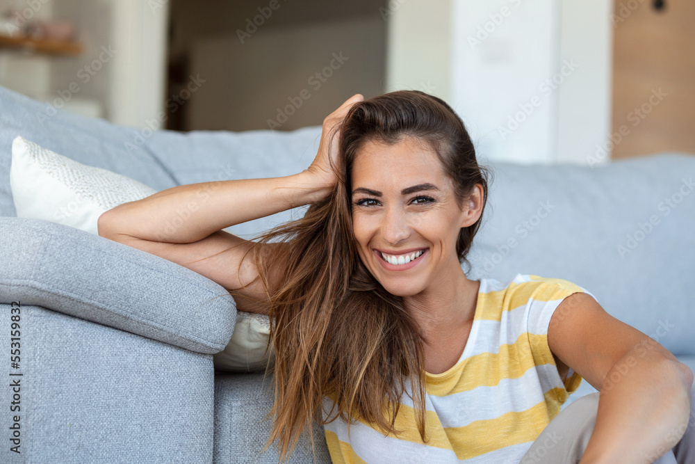 Naklejka premium Happy young woman sitting on sofa at home and looking at camera. Portrait of beautiful woman smiling and relaxing during summer.